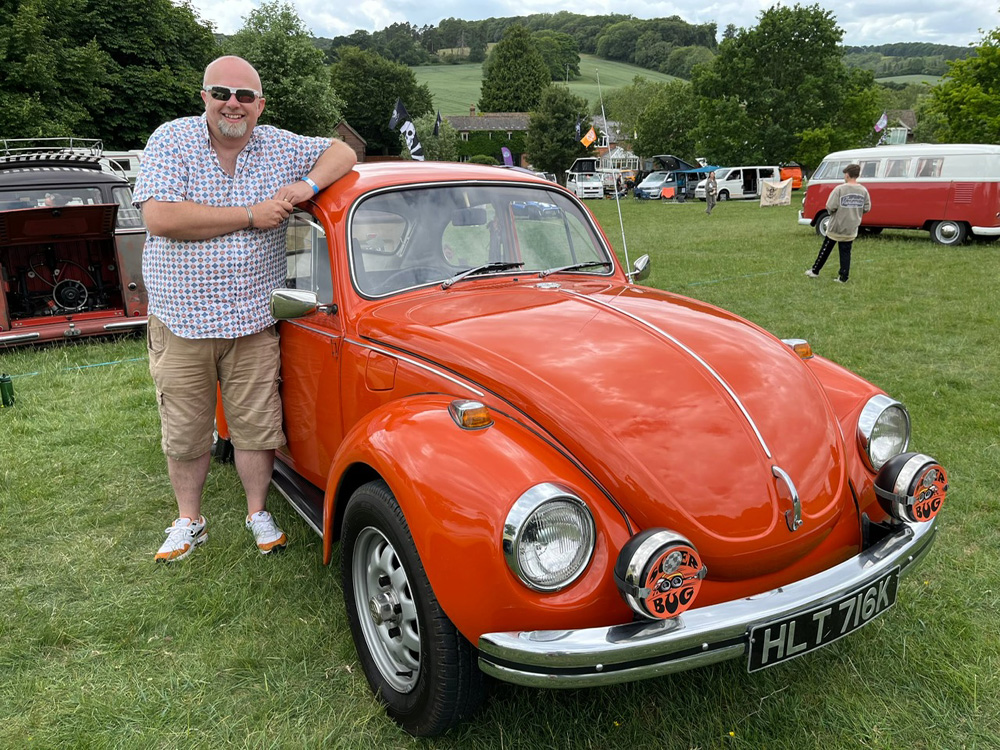 Mark and his Orange VW Super Beetle at an outdoor car show surrounded by hills and trees