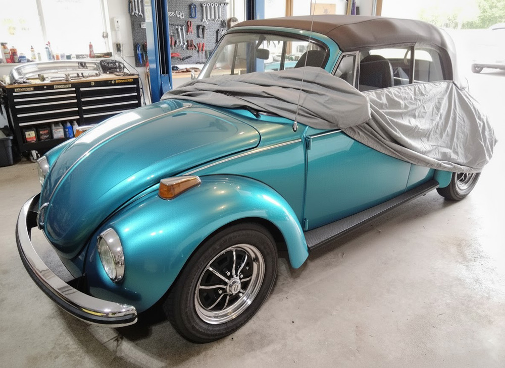 A teal vintage Super Beetle convertible Volkswagen being prepared for spring in a garage, partially covered by a protective car cover.