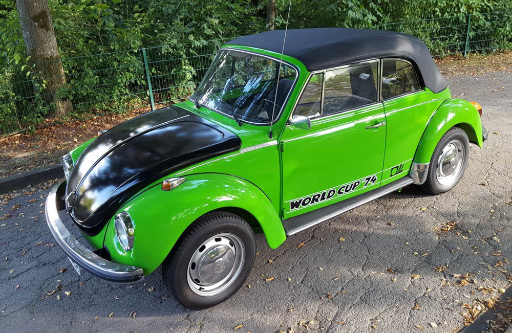 Side profile of a restored green 1974 World Cup Super Beetle convertible with black top and World Cup '74 graphics parked along a tree-lined road