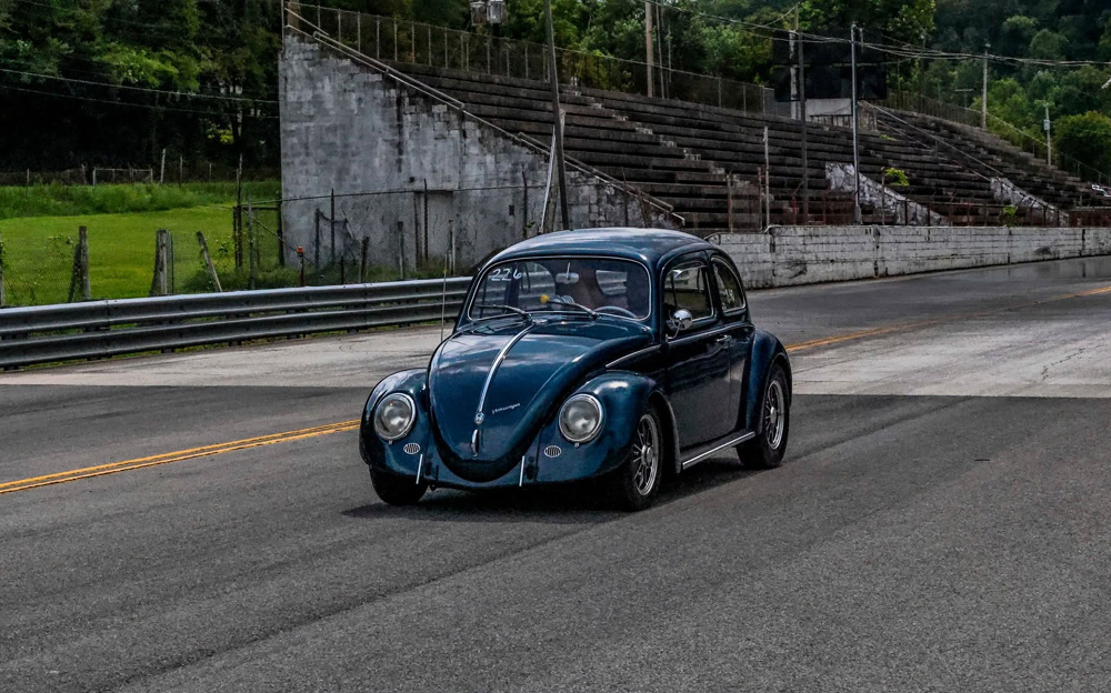 Dark blue VW Beetle driving past old concrete bleachers at a racetrack