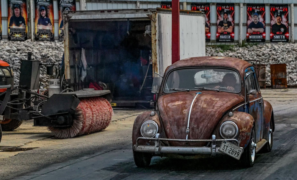 Weathered VW Beetle driving past posters and equipment at a motorsport venue